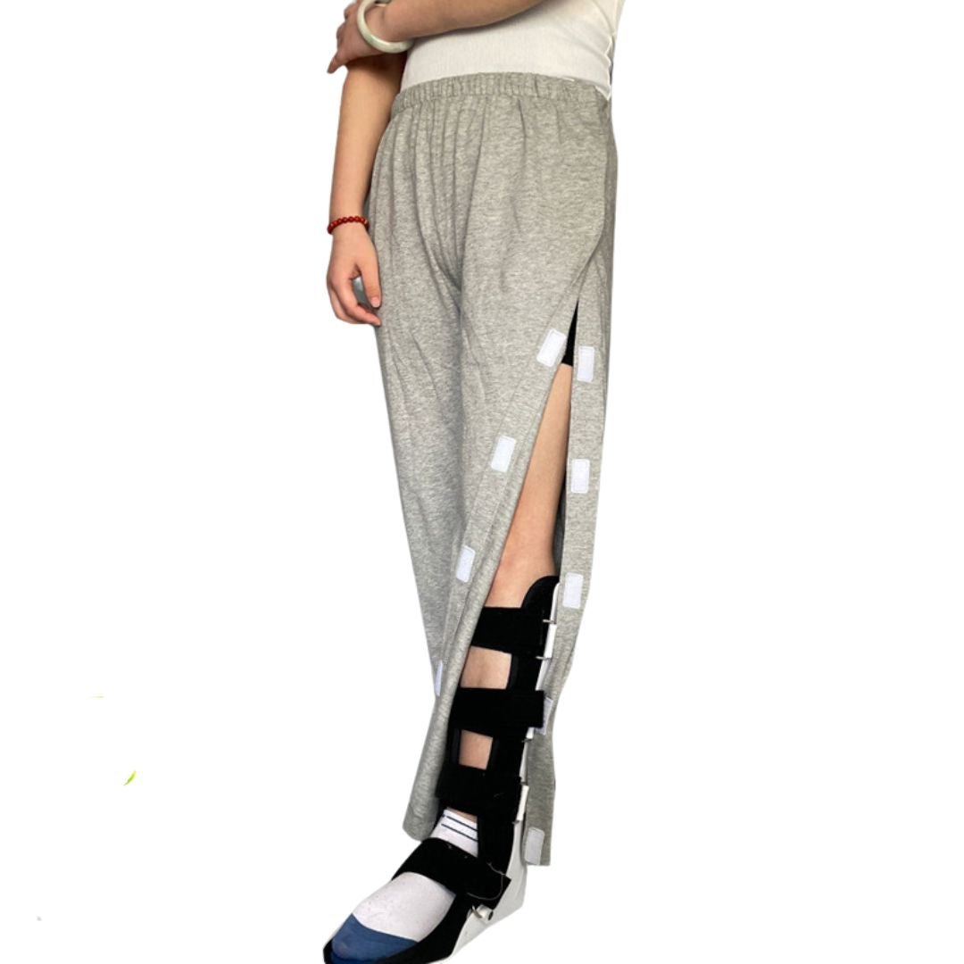 Woman standing wearing gray adaptive full open side pants with velcro closure, also visible is a supportive brace on her left leg.