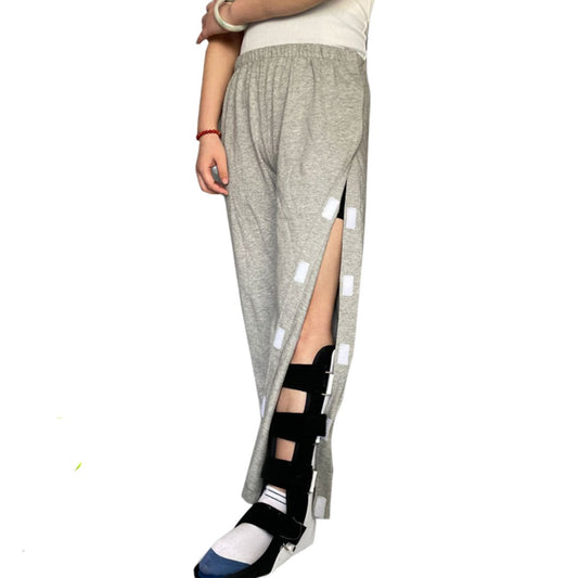 Woman standing wearing gray adaptive full open side pants with velcro closure, also visible is a supportive brace on her left leg.